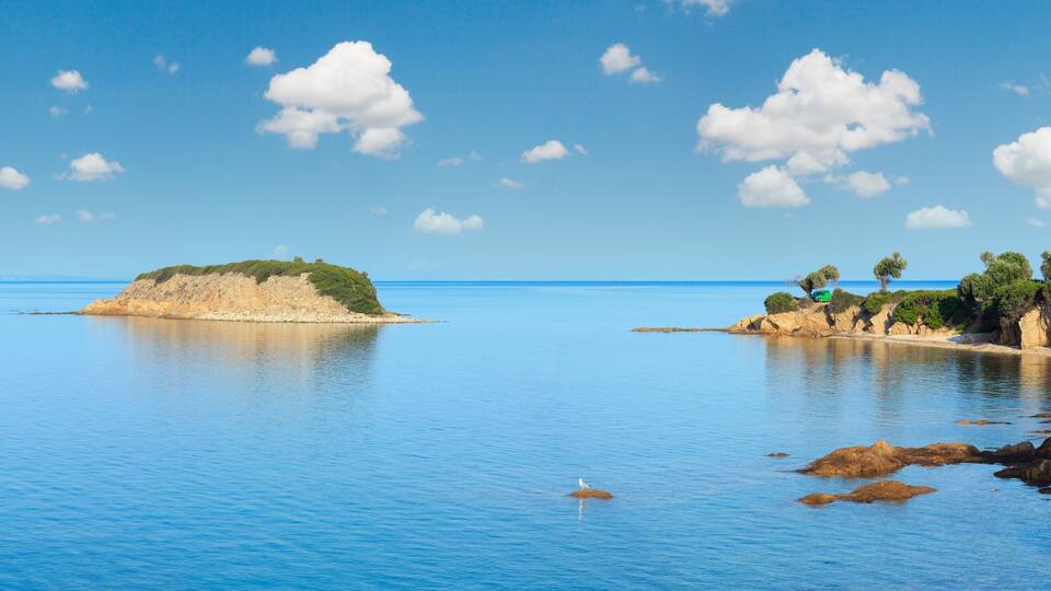 A calm sea with a small island, rocky coastline, and a clear blue sky.