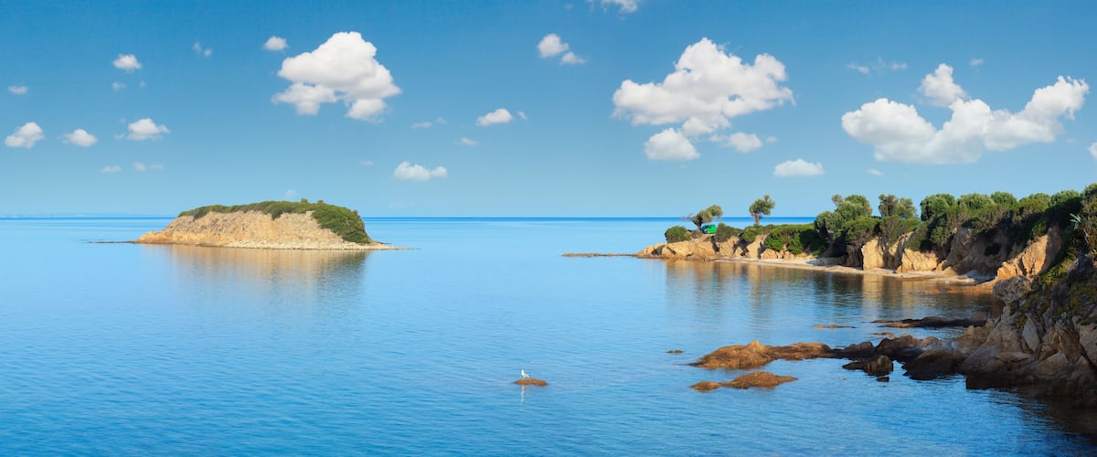 A calm sea with a small island, rocky coastline, and a clear blue sky.