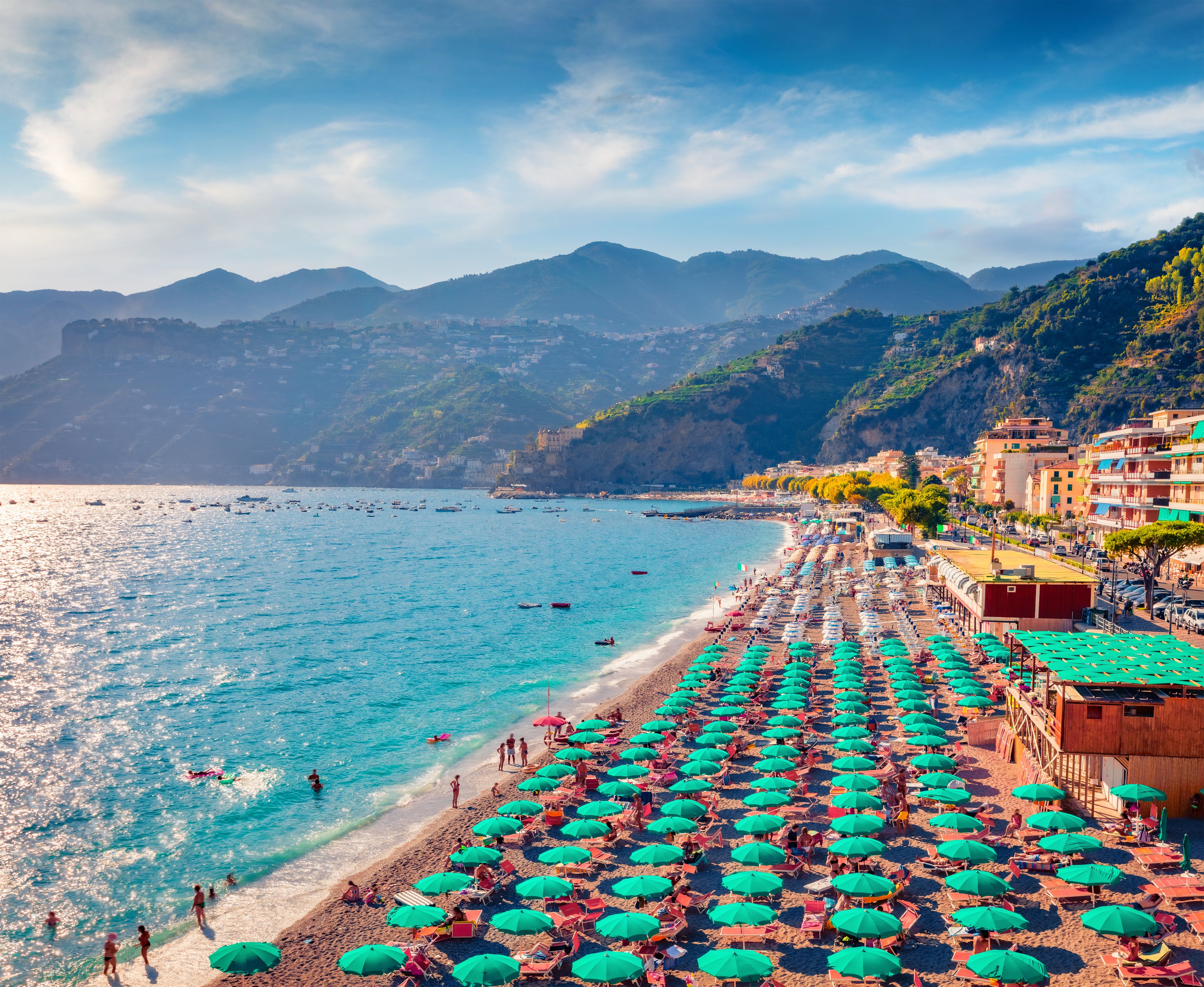 A coastal town with a beach lined with umbrellas, clear blue sea, and mountains in the background.