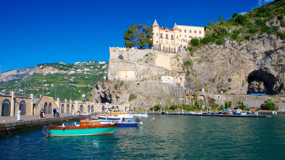 A coastal town with a historic castle on a cliff, boats docked at the harbor, and a rocky hillside in the background.