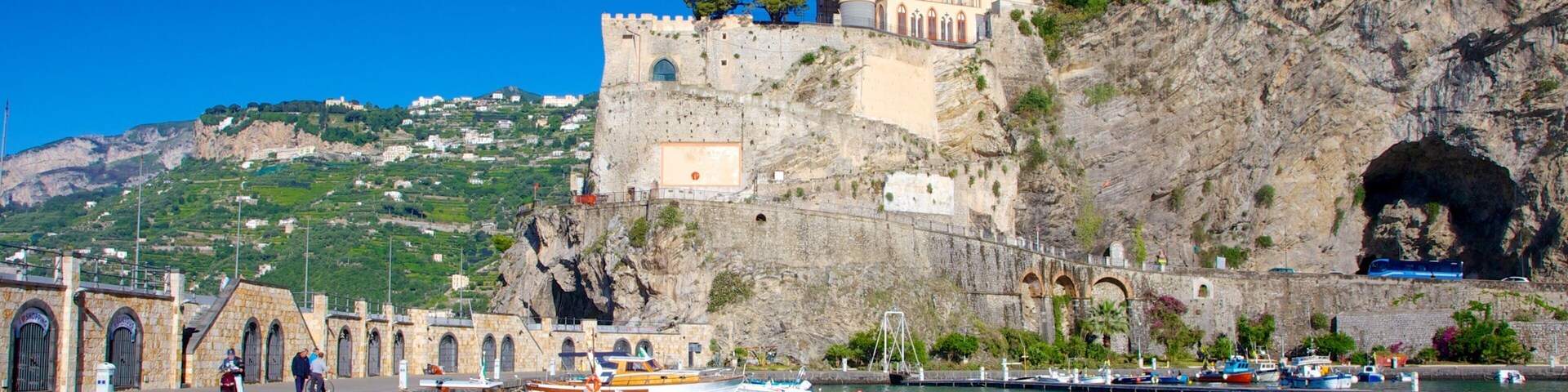 A coastal town with a historic castle on a cliff, boats docked at the harbor, and a rocky hillside in the background.