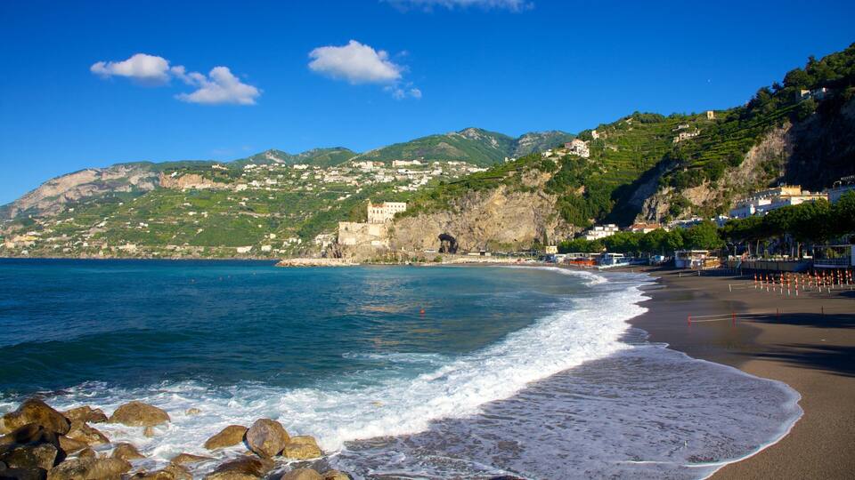 A coastal town with a beach, rocky shoreline, and a backdrop of green hills and buildings.