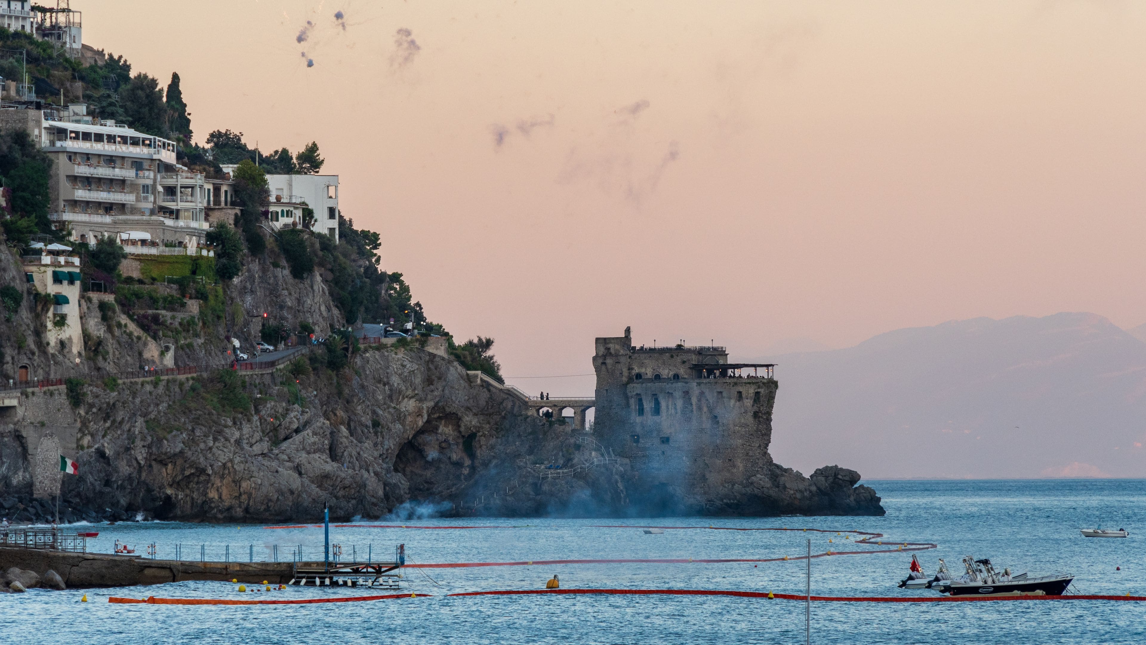 A coastal town with a historic castle on a cliff, boats in the water, and a clear sky.