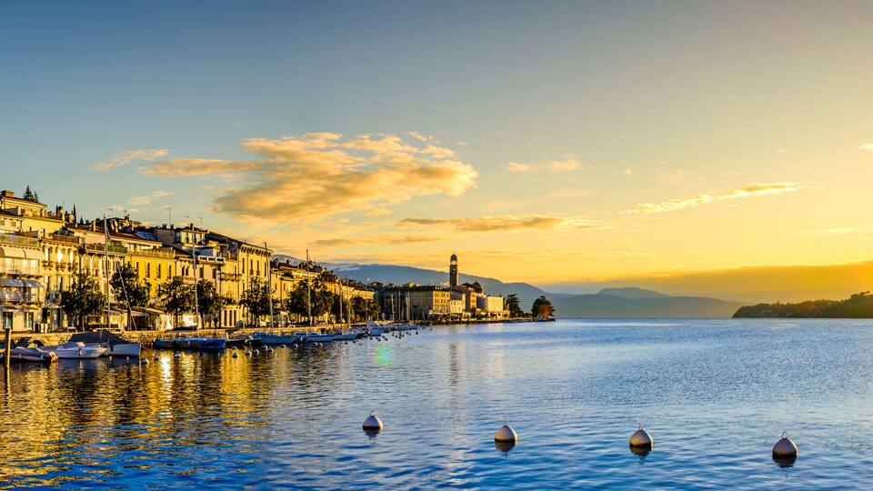 A waterfront cityscape at sunset with boats, buildings, and a hill in the background.