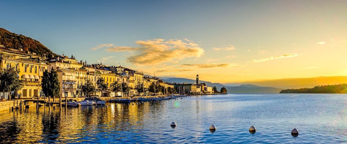 A waterfront cityscape at sunset with boats, buildings, and a hill in the background.