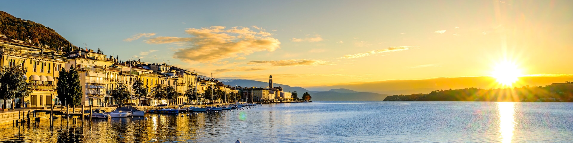 A waterfront cityscape at sunset with boats, buildings, and a hill in the background.