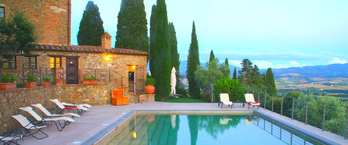 A swimming pool with lounge chairs, a stone building, and tall trees in the background.
