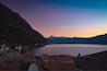 A lakeside town with red-roofed buildings, a mountain range, and a sunset sky.