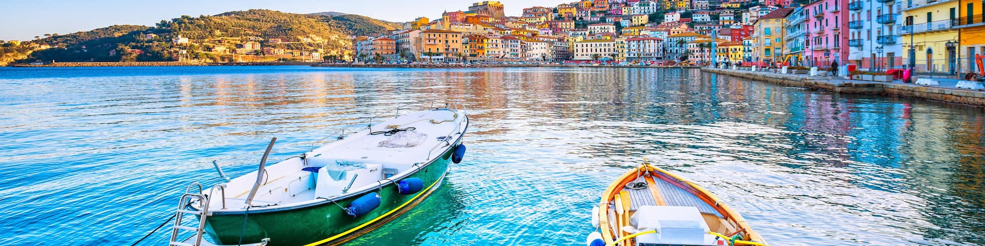 Two boats tied to the dock in a clear blue harbor with colorful buildings in the background.