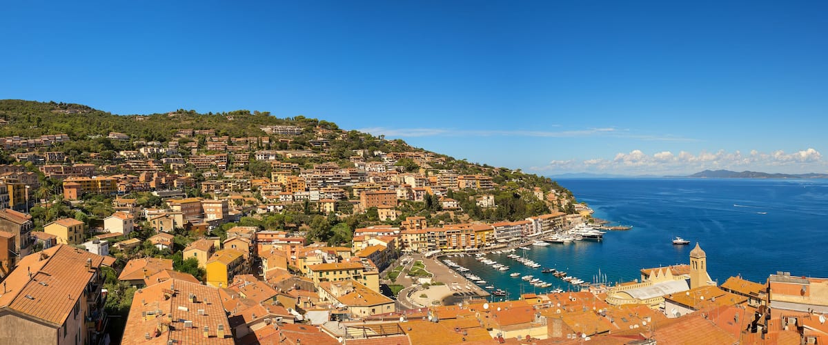 A coastal town with terracotta-roofed buildings, a marina, and a clear blue sea.