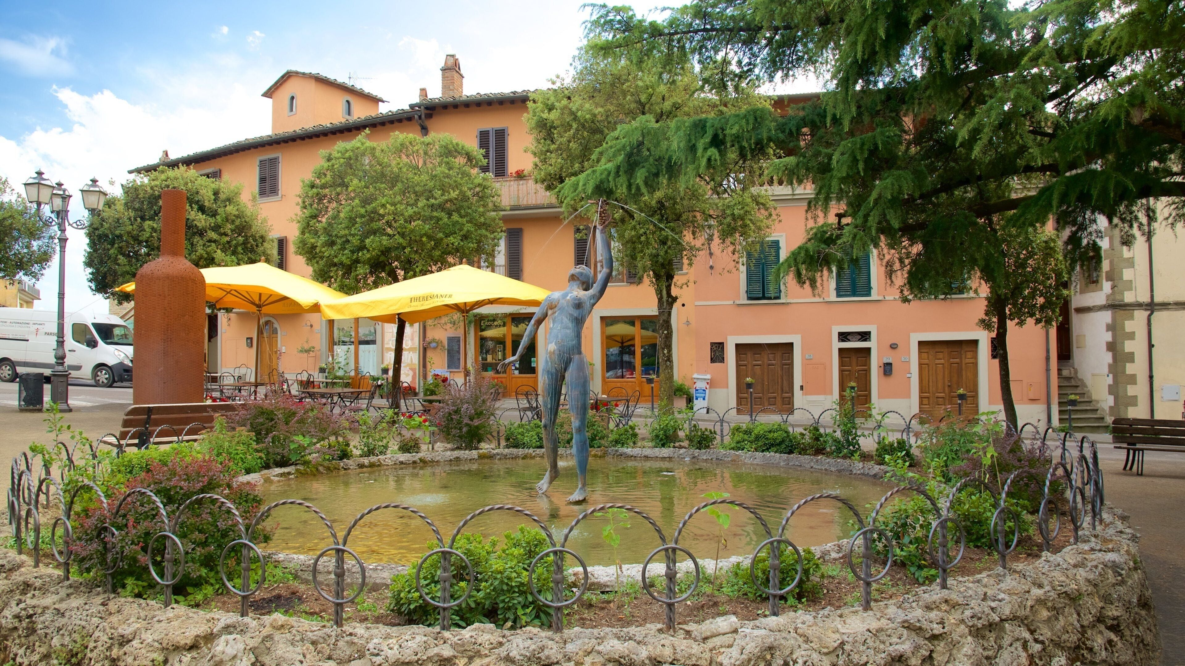 A fountain with a statue, surrounded by a stone wall and greenery, in front of a building with yellow umbrellas and outdoor seating.