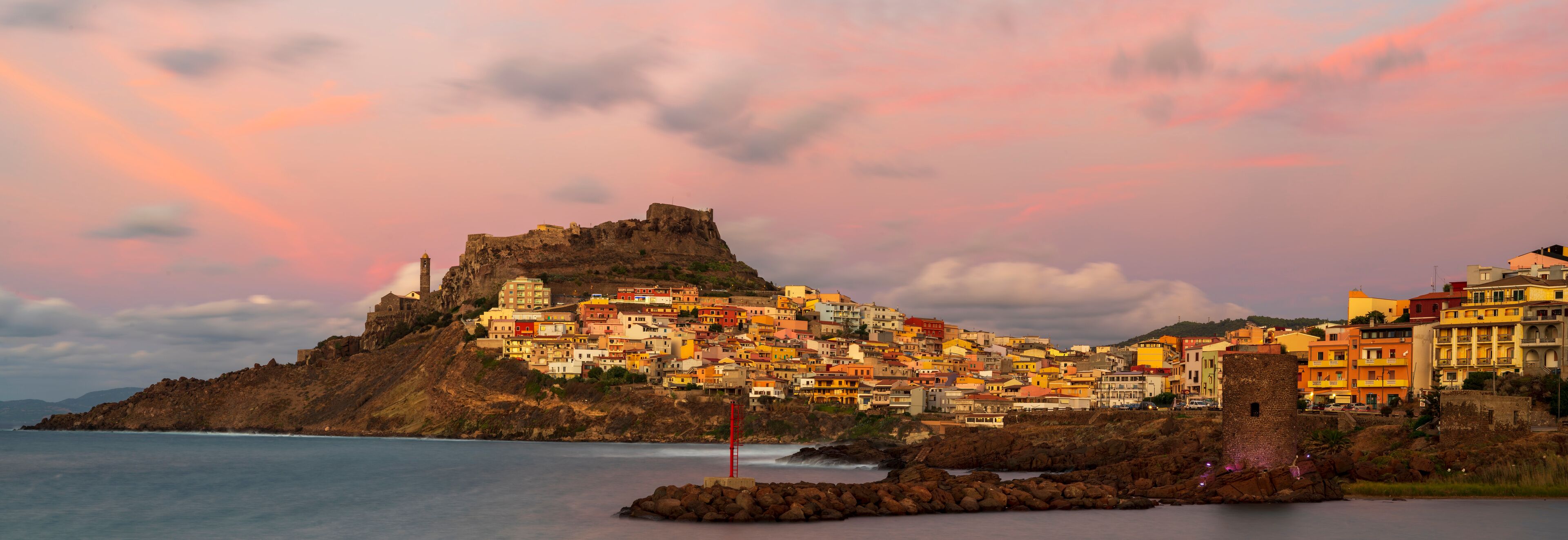 A coastal town with colorful buildings, a prominent castle on a hill, and a rocky shoreline.