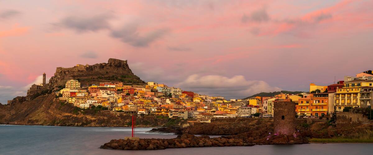 A coastal town with colorful buildings, a prominent castle on a hill, and a rocky shoreline.