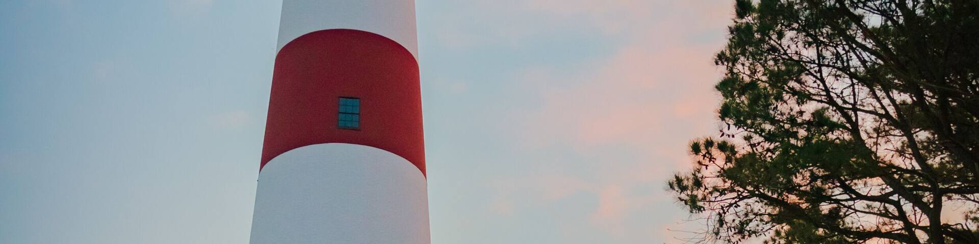 A tall red and white lighthouse with a small red building at its base, surrounded by trees and a gravel path.