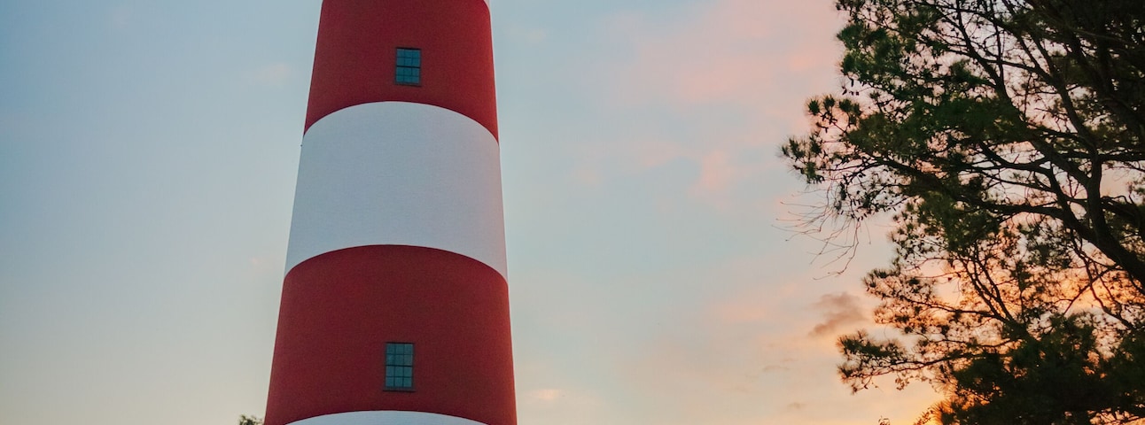 A tall red and white lighthouse with a small red building at its base, surrounded by trees and a gravel path.
