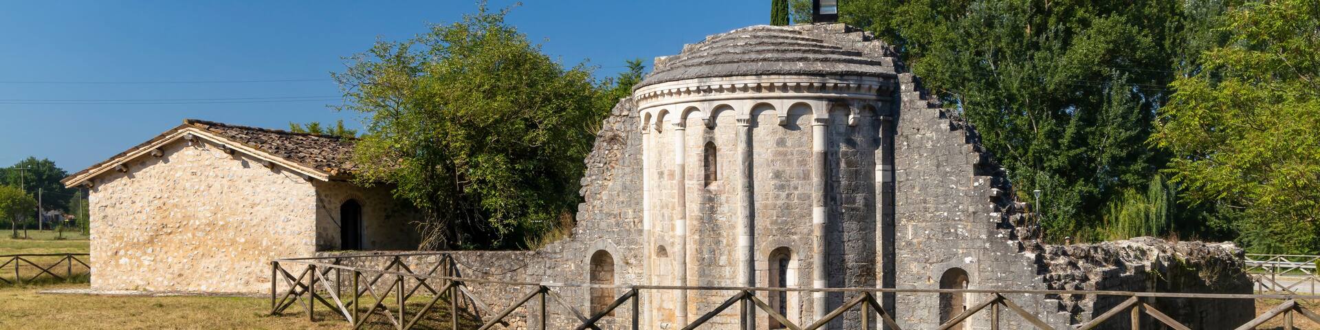 An ancient stone building with a dome and arches, surrounded by a fenced area and trees.
