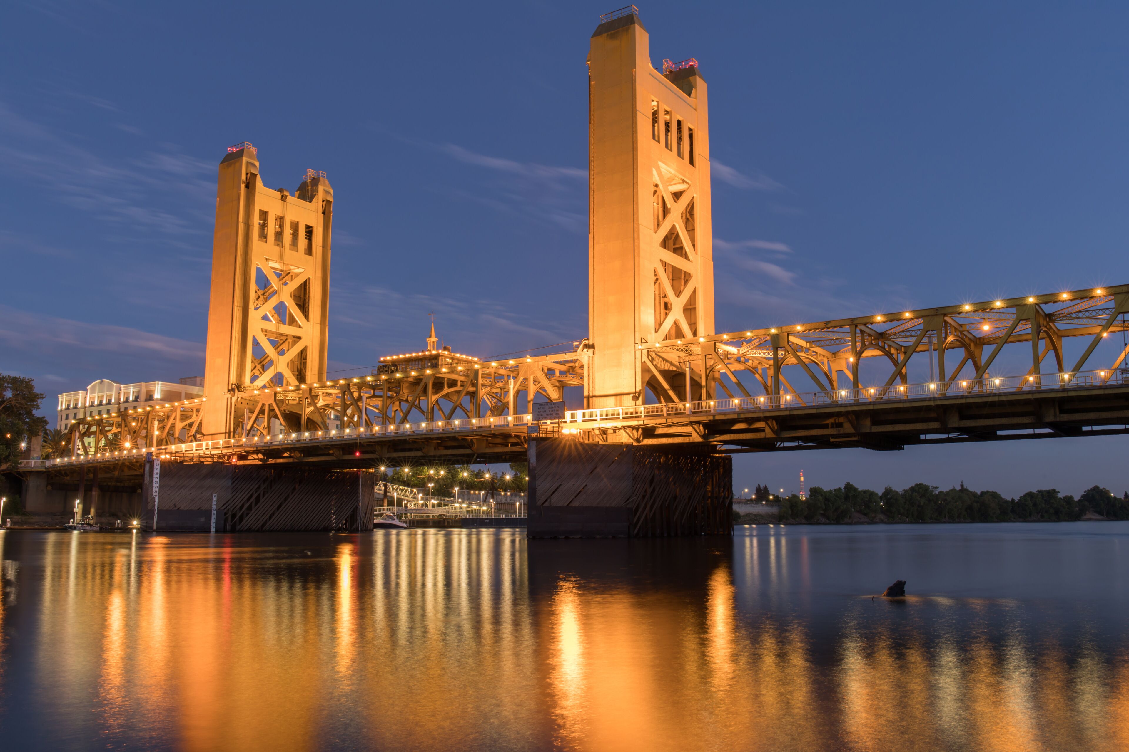 A large steel bridge spanning a river at dusk with lights reflecting on the water.