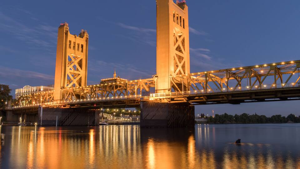 A large steel bridge spanning a river at dusk with lights reflecting on the water.