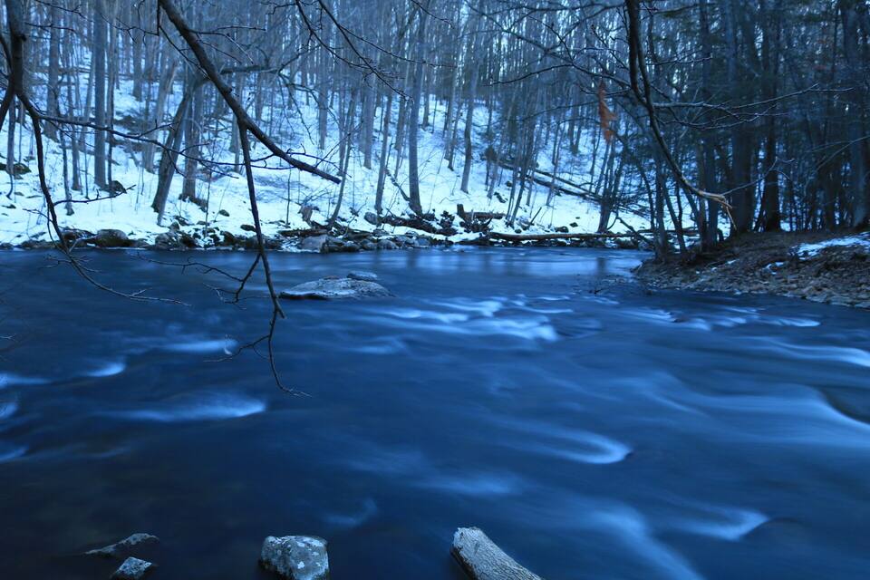 A frozen river in a forest with bare trees and snow on the ground.