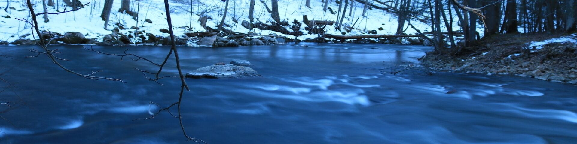 A frozen river in a forest with bare trees and snow on the ground.