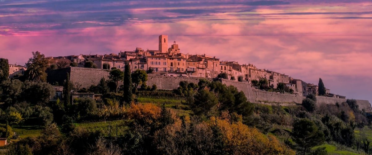 A historic town perched on a hill with a prominent tower, surrounded by lush greenery and a dramatic sunset sky.
