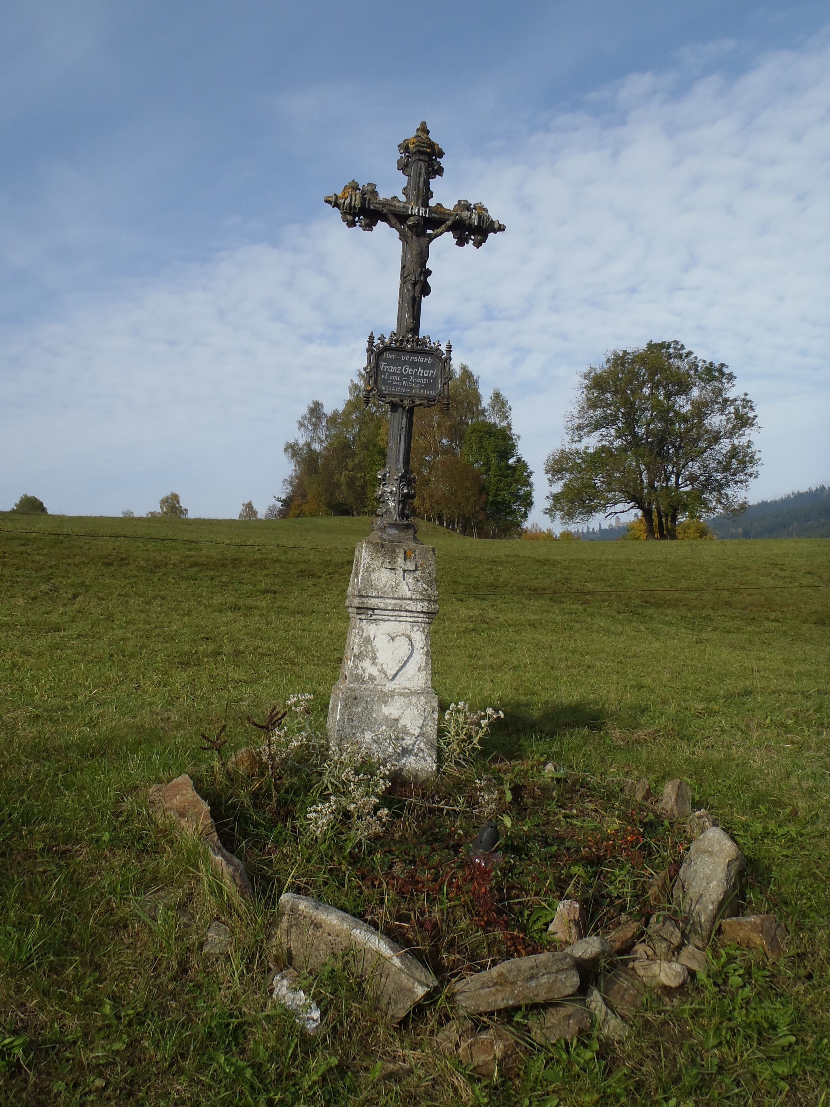 Uma cruz de pedra desgastada com inscrições em um campo gramado, cercada por pedras e árvores.