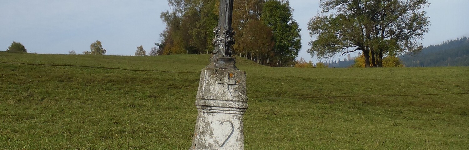 Uma cruz de pedra desgastada com inscrições em um campo gramado, cercada por pedras e árvores.