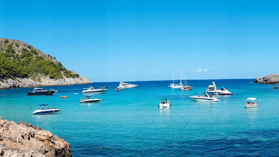 A coastal bay with clear turquoise waters, rocky shores, and a forested hill.