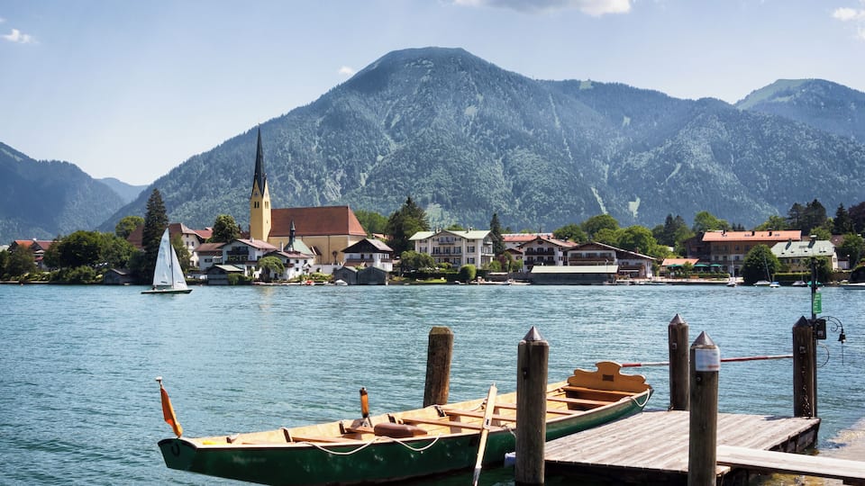 A lake with a sailboat docked at a wooden pier, surrounded by houses and a church with a tall steeple.
