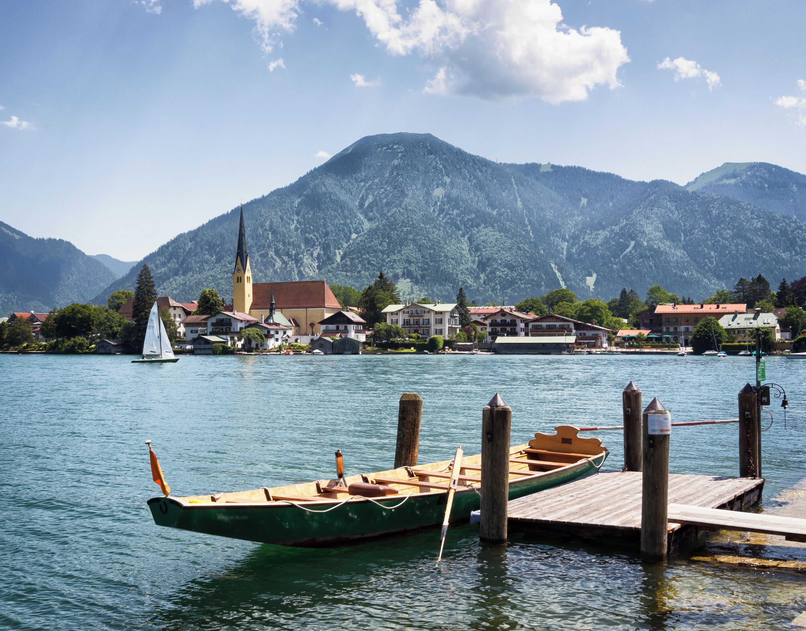 A lake with a sailboat docked at a wooden pier, surrounded by houses and a church with a tall steeple.