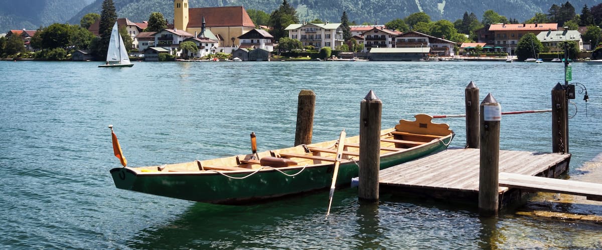 A lake with a sailboat docked at a wooden pier, surrounded by houses and a church with a tall steeple.