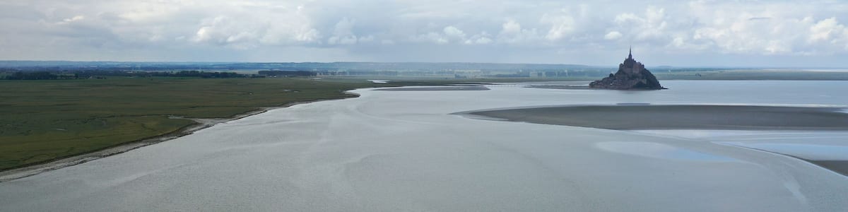 Eine Küstenlandschaft mit einem großen Gewässer, einer kleinen Insel mit einer festungsähnlichen Struktur und sanften Hügeln unter bewölktem Himmel.