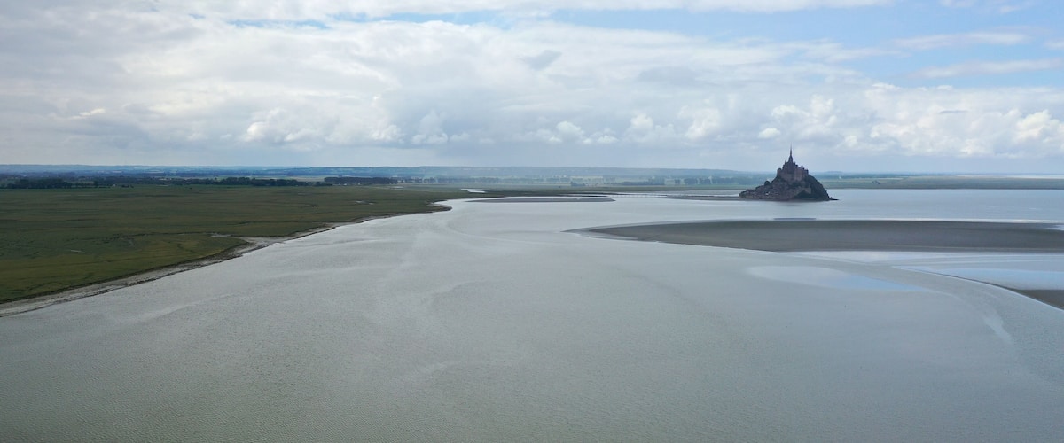 Un paysage côtier avec une vaste étendue d’eau, une petite île surmontée d’une structure ressemblant à un château, et des collines lointaines sous un ciel nuageux.