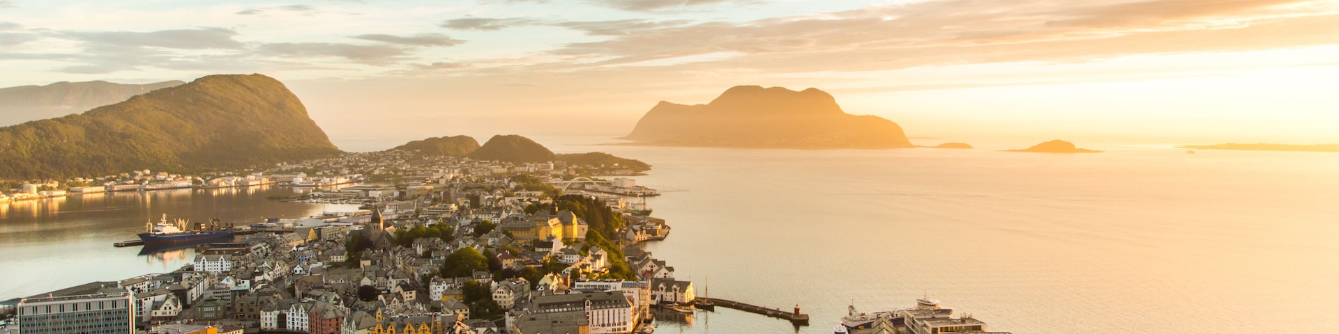 Un paisaje urbano costero con un puerto destacado, edificios históricos y montañas al fondo durante el atardecer.