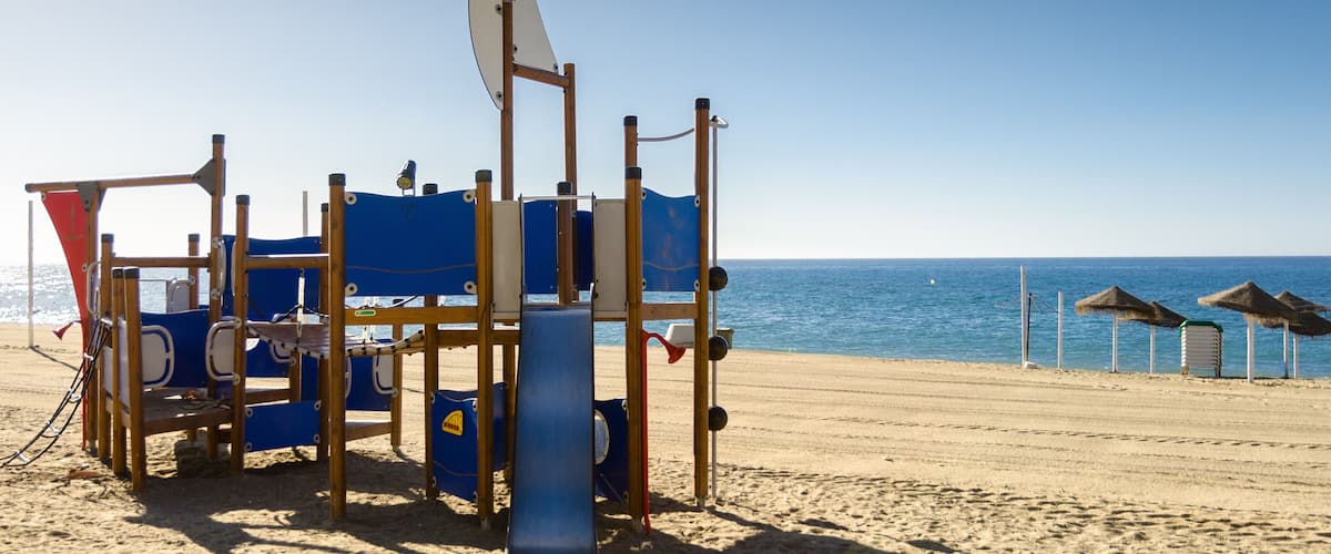 A beach with a children's play structure, a flag, and thatched umbrellas.