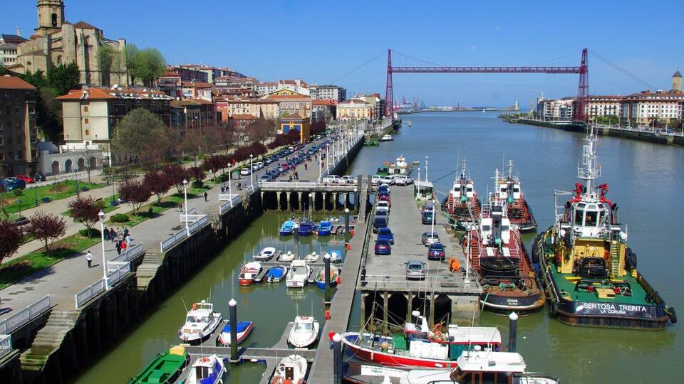 A harbor with boats docked, a bridge in the background, and a historic building.