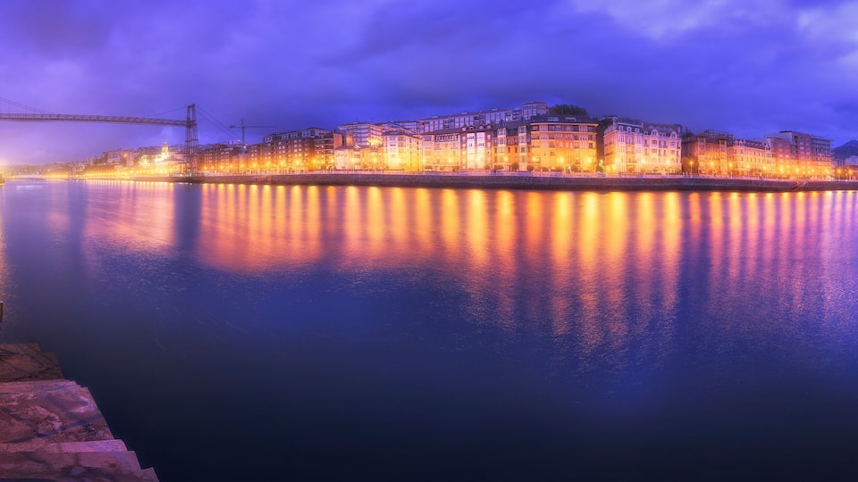 A waterfront cityscape at dusk with illuminated buildings reflecting on the water.