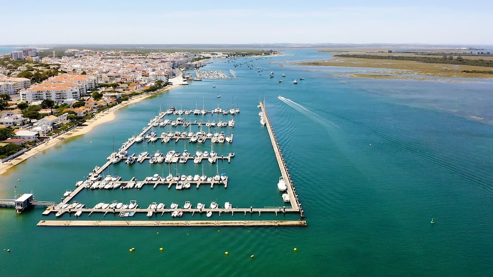 An aerial view of a marina with numerous boats docked, a long pier extending into the water, and a coastal cityscape in the background.