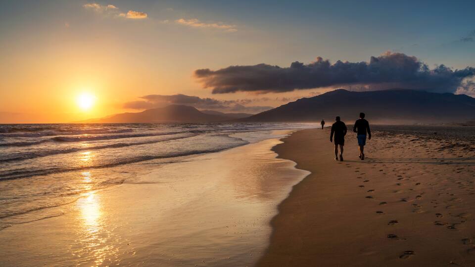 A beach with two people walking, the sun setting over the ocean, and mountains in the background.