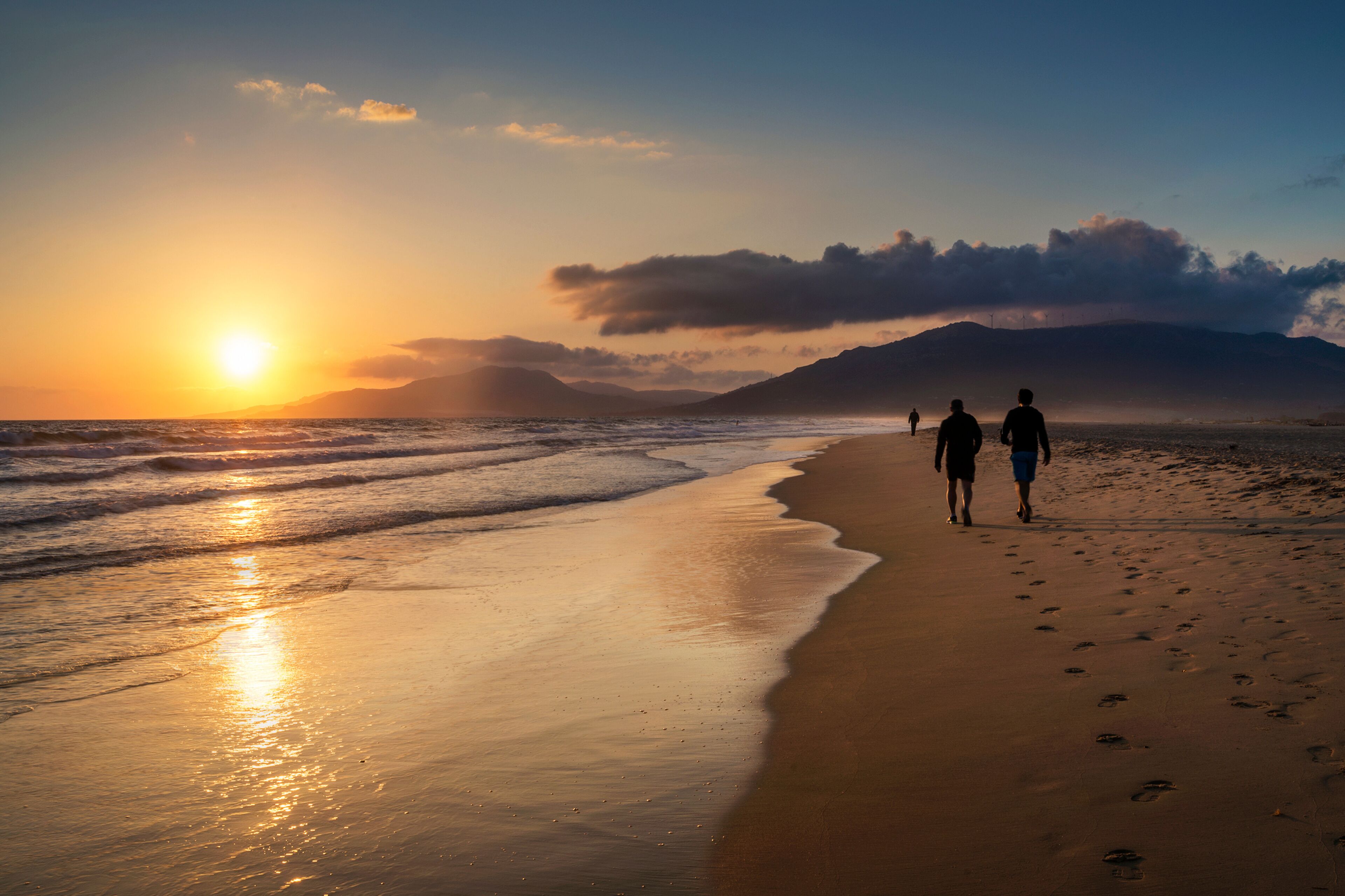 A beach with two people walking, the sun setting over the ocean, and mountains in the background.