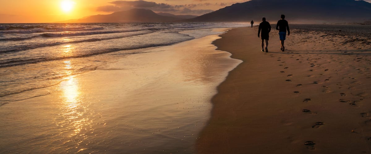 A beach with two people walking, the sun setting over the ocean, and mountains in the background.
