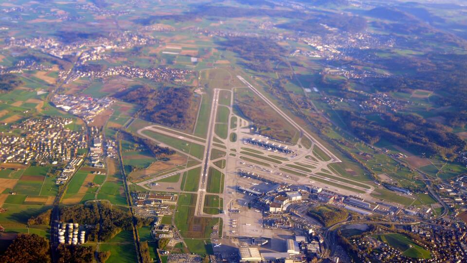 An aerial view of a complex airport layout with multiple runways and taxiways, surrounded by agricultural fields and residential areas.