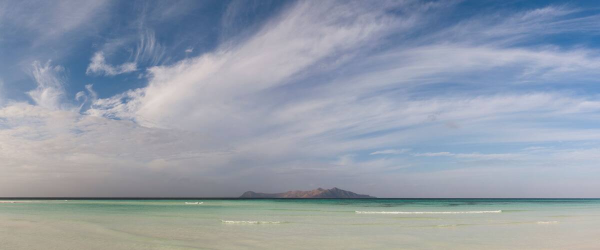 A beach with clear turquoise water, white sand, and distant mountains under a partly cloudy sky.