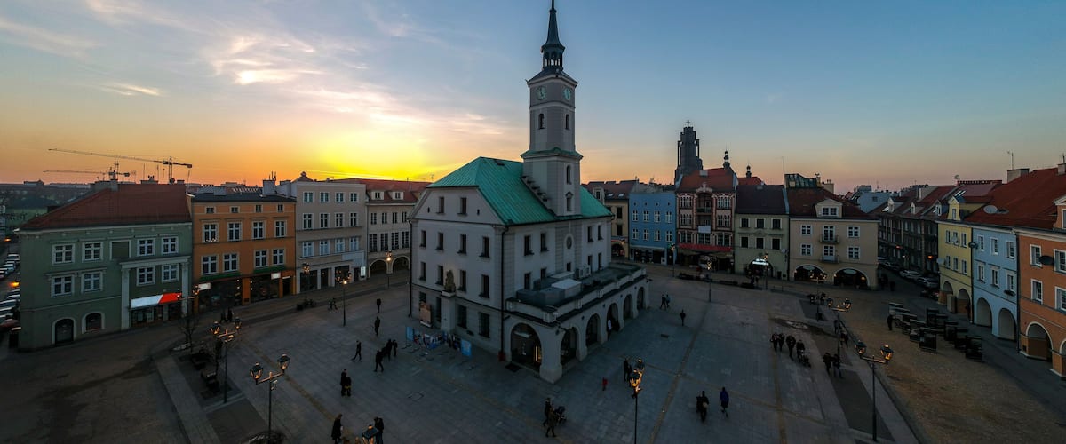 A city square with a prominent church tower, historic buildings, and a sunset in the background.