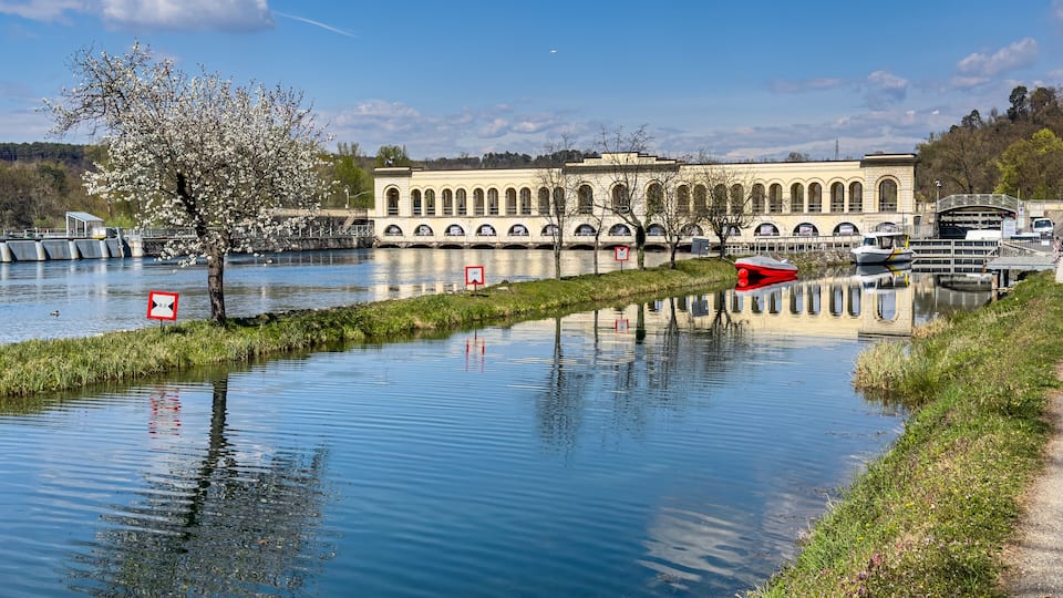 A calm river with a bridge, a tree with white blossoms, and a building with arched windows in the background.