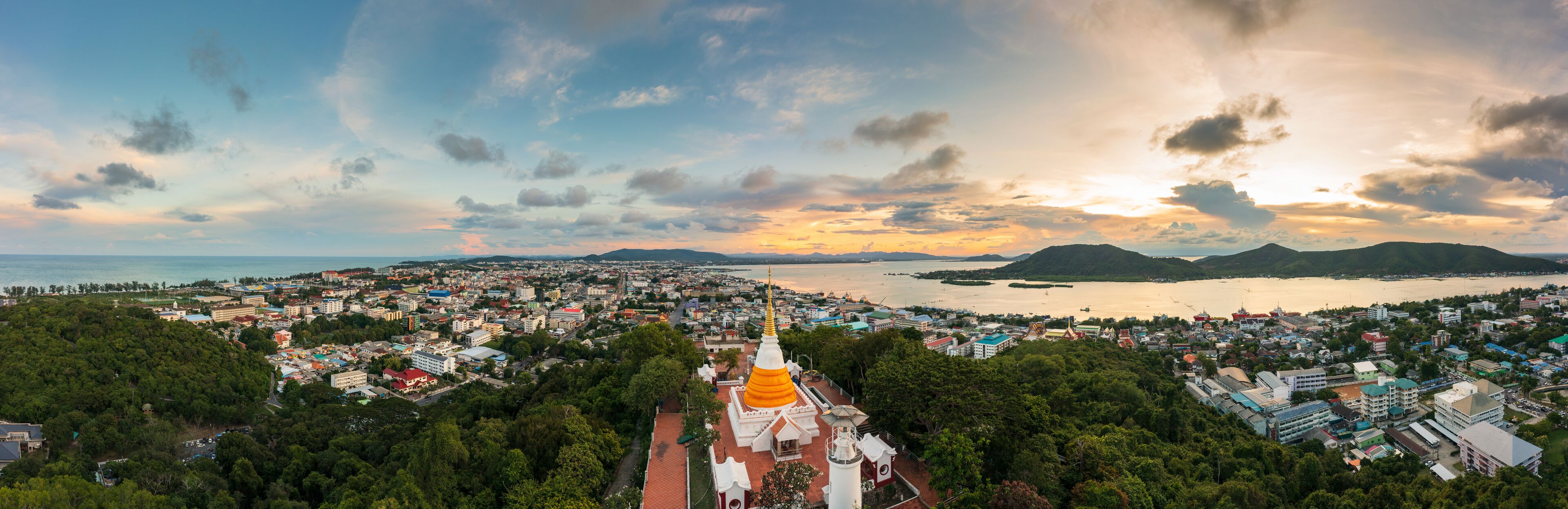 A coastal cityscape with a prominent golden-roofed temple, lush greenery, and a sunset over the water.