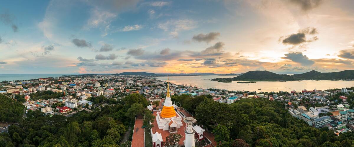 A coastal cityscape with a prominent golden-roofed temple, lush greenery, and a sunset over the water.
