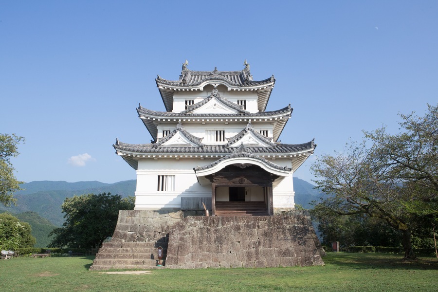 Un castillo japonés tradicional con varios niveles y base de piedra, rodeado de césped y árboles.