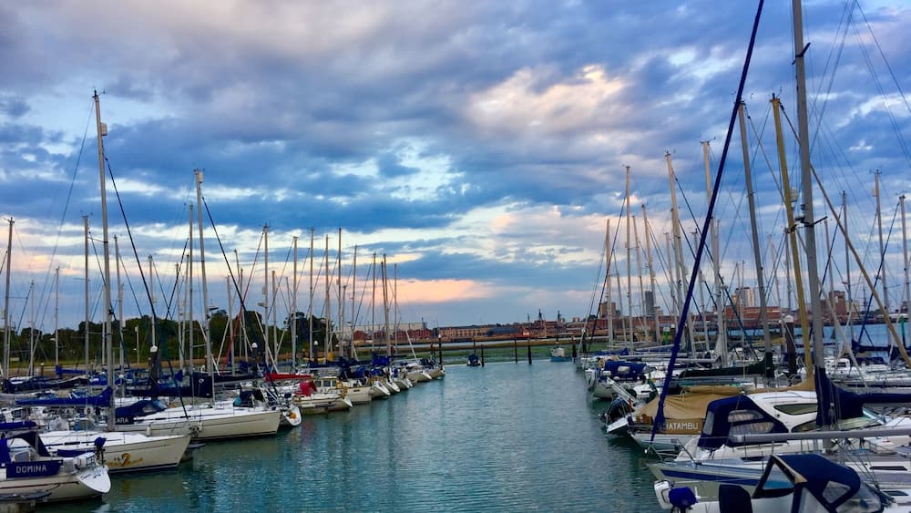 A marina with numerous sailboats docked along a calm waterway, under a partly cloudy sky.