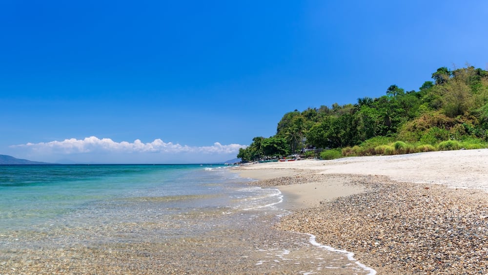 Une plage de galets bordée d’une eau turquoise et limpide, une étendue de sable et, en arrière-plan, une colline boisée.